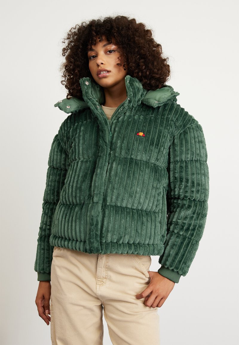 Groene pufferjack met ribbeltextuur, voorzien van een ritssluiting, capuchon en kleurrijk logo op de borst. G gedragen met lichtbeige broeken.