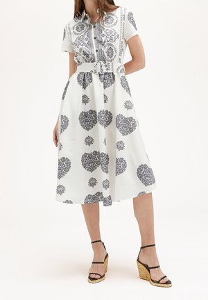 Femme portant une robe midi blanche ceinturée avec des motifs noirs complexes en forme de cœur et de fleurs, associée à des sandales compensées noires à lanières.