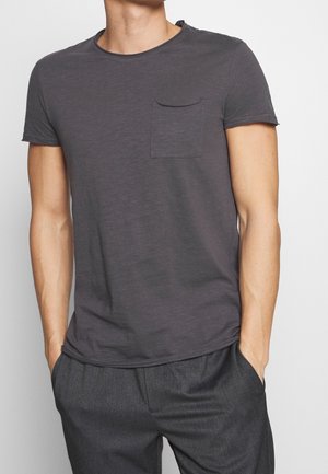 Mann trägt ein dunkelgraues kurzärmeliges T-Shirt mit Brusttasche und dunkelgraue Hose, steht mit Händen in den Taschen.