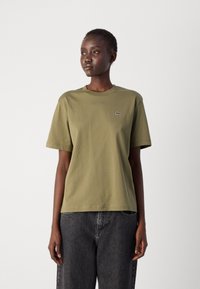 Olivgrünes Baumwoll-T-Shirt mit kurzen Ärmeln, Rundhalsausschnitt und kleinem gesticktem Logo auf der Brust, kombiniert mit schwarzen Denim-Hosen.