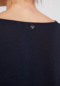 Takana näkymä henkilöstä, joka wear a tummansininen paita, jossa on pieni pyöreä hopeinen merkki kaulan yläpuolella, jossa lukee "rich&royal."