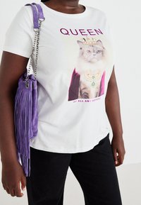 Person som bär en vit t-shirt med en katt som bär en krona och halsband, texten "QUEEN of all and nothing", och en lila väskan med fransar på axeln.