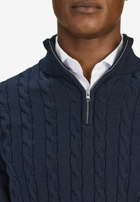 Marinblå kabelstickad tröja med hög krage, med en dragkedjeslutning. Under tröjan syns en vit skjorta med krage vid halsen.