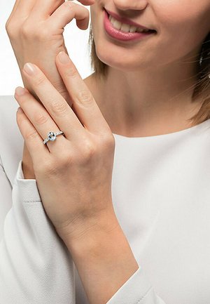 Silberring mit einem ovalen blauen Edelstein, umgeben von kleineren klaren Steinen. Die Hand ruht auf einem hellen Stoffhintergrund.