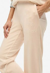 Beige vrijetijdspantalons met geborduurd "Superdry" logo op de bovenbeen, gedragen met één hand in de zak en de andere langs het lichaam.