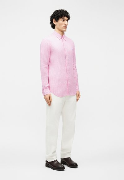 Άνδρας που στέκεται, φοράει ροζ πουκάμισο με μακριά μανίκια και κουμπιά, λευκό παντελόνι και σκούρα καφέ παπούτσια, κοιτάζοντας ελαφρώς προς τα δεξιά σε λευκό φόντο.