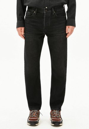 Person trägt schwarze Jeans, schwarzes Hemd und braune Sportschuhe mit grauen Schnürsenkeln und steht vor weißem Hintergrund.