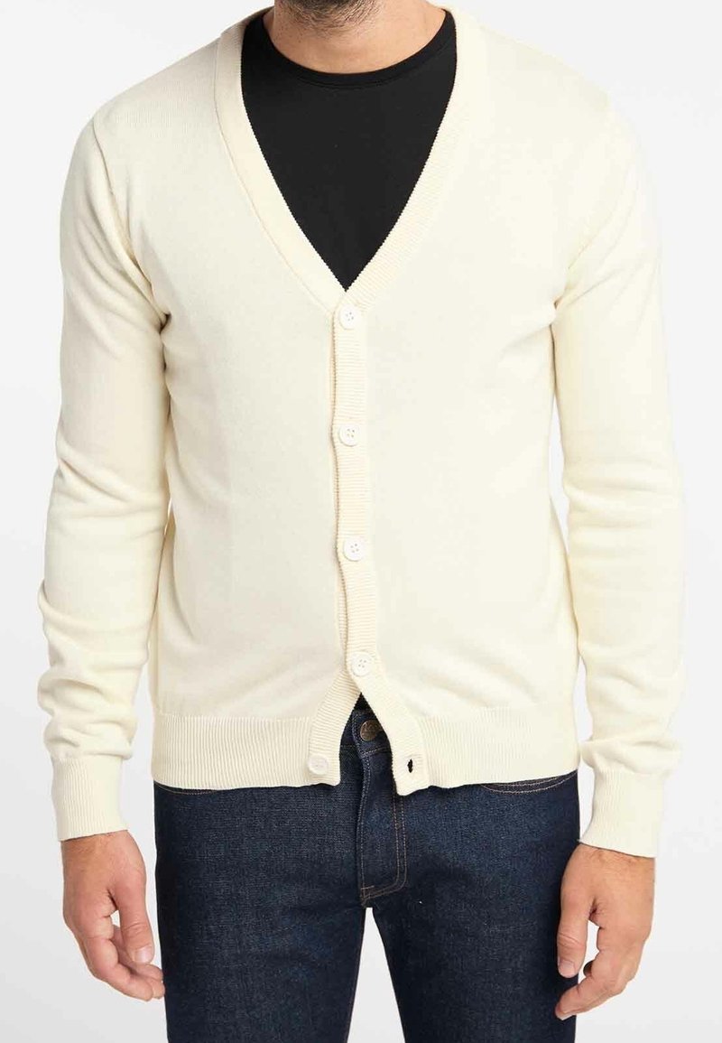 Cremefarbener Cardigan mit V-Ausschnitt, Knopfverschluss, gerippten Ärmelbündchen und Saum, aus weichem Material, getragen über einem schwarzen Hemd.