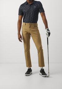 Man wearing navy patterned polo, tan pants, black golf shoes, and a white glove holding a golf club, standing on a plain white background.