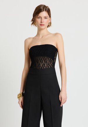 Donna con top nero senza spalline in pizzo e pantaloni neri a vita alta e gamba larga, accessoriata con orecchini a cerchio dorati e numerosi braccialetti d'oro.