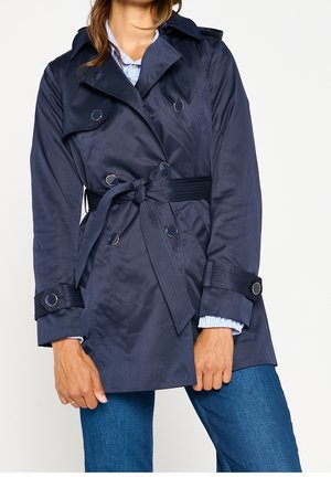 Vrouw die een marineblauwe trenchcoat met ceintuur en zilveren knopen draagt, over een blauwe spijkerbroek en met zichtbare boordjes van een gestreept shirt.