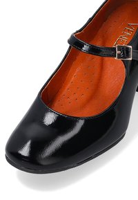 Black patent leather Mary Jane shoe with a round toe, ankle strap, and orange insole featuring perforations for breathability.
