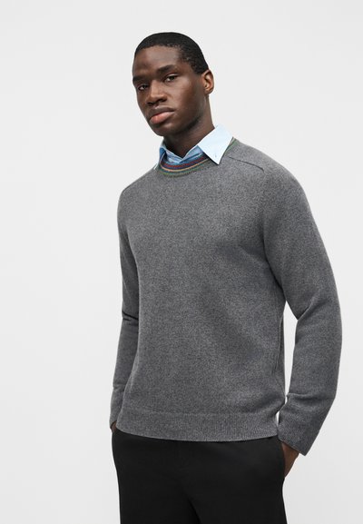 Jeune homme les mains dans les poches, portant un pull gris par-dessus une chemise bleue à col, sur un fond clair uni.