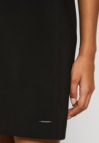 Sorte nederdel lavet af glat stof, med en sidesøm og et metal logo-tag i kanten. Designet er enkelt uden mønstre.