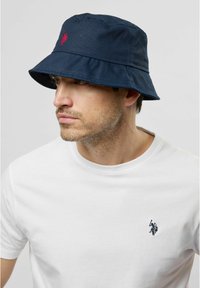 Marinblå bucket hat med bred skärm och en röd broderad logga. Bärs över en vit t-shirt med en liten marinblå logga. Tillverkad av bomull.