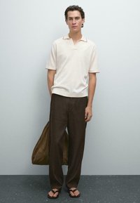 Polo beige a maniche corte con colletto a contrasto, abbinato a pantaloni larghi in lino marrone e ciabatte nere, mentre tiene in mano una borsa in suede marrone.