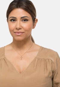 Beige bluse med v-hals, med samlet detaljering. Gylden halskjede med et korsanheng. Hudtonen er varm, med synlig glatt tekstur.