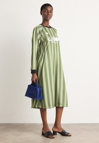 Ein olivgrünes gestreiftes Kleid mit schwarzen Akzenten und einem Logo. Das Model hält eine kleine blaue Handtasche und trägt schwarze Slipper.