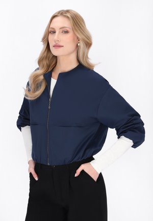 Kvinde iført marineblå lynlåstrøje over hvide langærmer og sorte bukser, hænderne i lommerne, poserer mod en ensfarvet hvid baggrund.