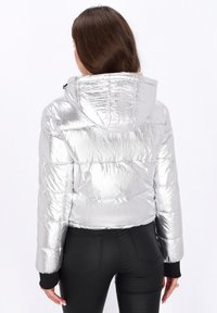 Giacca puffer argentea con cappuccio, caratterizzata da una superficie lucida e testurizzata, polsini elasticizzati e un design cropped. Pantaloni neri visibili sotto.