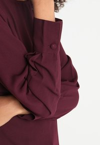 Chemise bordeaux à manches longues avec poignet boutonné, texture douce et détails froncés près du poignet pour une coupe décontractée.