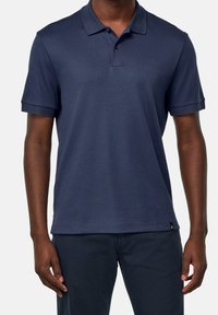 Námořnická modrá polo košile vyrobená z hladké tkaniny. Má klasický límec, krátké rukávy a zapínání na tři knoflíky. Diskrétní štítek značky na lemu.
