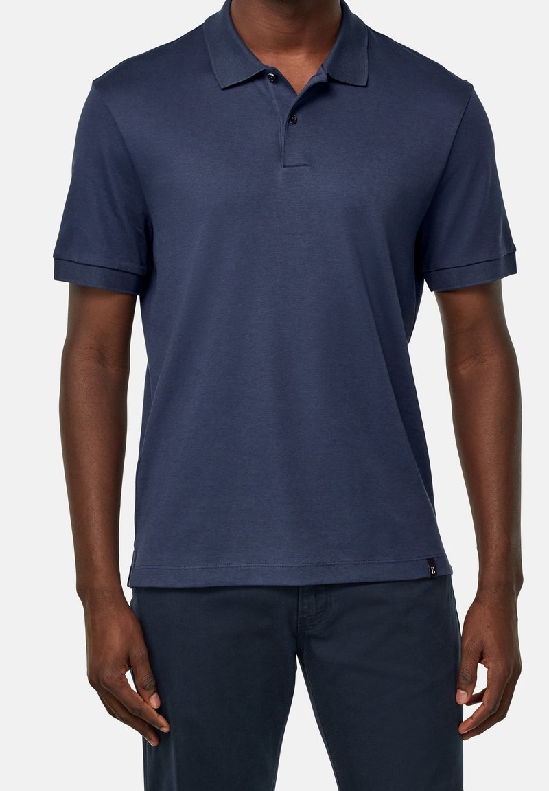 Námořnická modrá polo košile vyrobená z hladké tkaniny. Má klasický límec, krátké rukávy a zapínání na tři knoflíky. Diskrétní štítek značky na lemu.