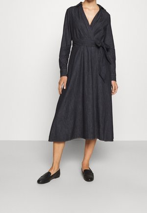 Frau trägt ein langärmliges, dunkles Wickelkleid mit gebundenem Gürtel, kombiniert mit schwarzen Slippern, stehend vor einem schlichten Hintergrund.