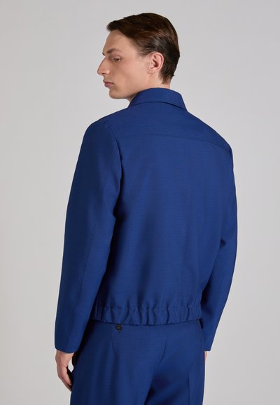 Man wearing a tailored, royal blue jacket and matching trousers, facing sideways with head turned slightly back against a plain background.