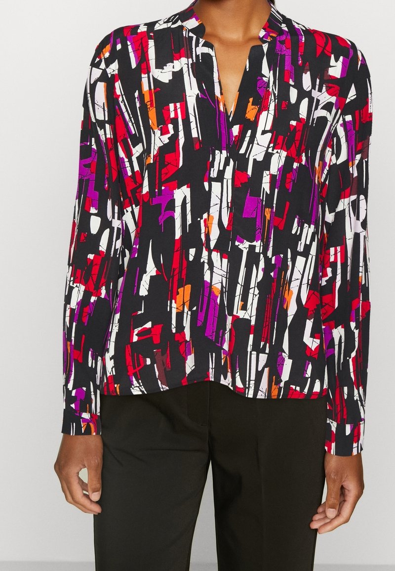 Personne portant un chemisier à manches longues à fond noir et imprimé abstrait audacieux rouge, blanc, violet et orange, assorti à un pantalon noir.