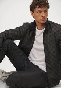 Giacca nera a motivi con zip frontale, abbinata a una camicia bianca e pantaloni neri. La giacca presenta una texture geometrica e un colletto alto.