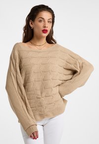 Pulover bej cu tricotaj tip cable-knit, cu o croială lejeră, decolteu larg și mansete cu nervuri, asortat cu pantaloni albi. Un model subtil de textură pe toată suprafața.