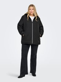 Blonde woman stands wearing a black zip-up puffer jacket, black flared pants, black boots, and a white top, hands in jacket pockets.