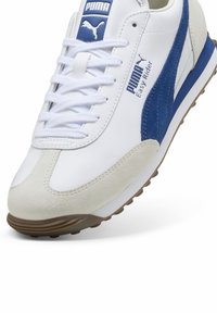 Les sneakers Puma Easy Rider présentent un dessus en cuir blanc avec des accents en suède bleu et un capuchon de pointe en suède gris, le tout complété par des lacets blancs et une semelle brune.