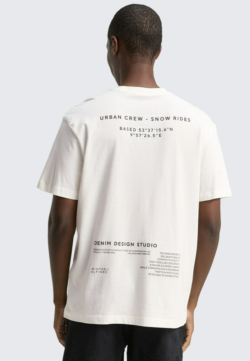 Witte katoenen t-shirt met ronde hals, voorzien van zwarte tekst op de achterkant met stedelijke grafische elementen en coördinaten. Relaxte pasvorm; korte mouwen.