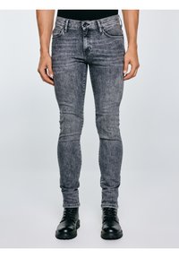 Mees, kes kannab kitsaid tuhmunud halli teksapükse ja musti paeltega saapaid, seisab ühevärvilise heleda tausta ees.