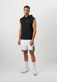 Sort ærmeløs hættebluse med kængurulomme og hvid Jordan-logo, parret med hvide mesh shorts med sorte striber og accenter.