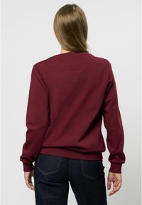 Felpa bordeaux con maniche lunghe e scollatura tonda, realizzata in tessuto morbido, presenta polsini e orlo a coste, abbinata a jeans scuri.