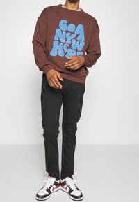 Uomo che indossa un felpa marrone con grandi lettere in stile uncinetto blu, pantaloni neri e sneakers bianche, nere e rosse in piedi.