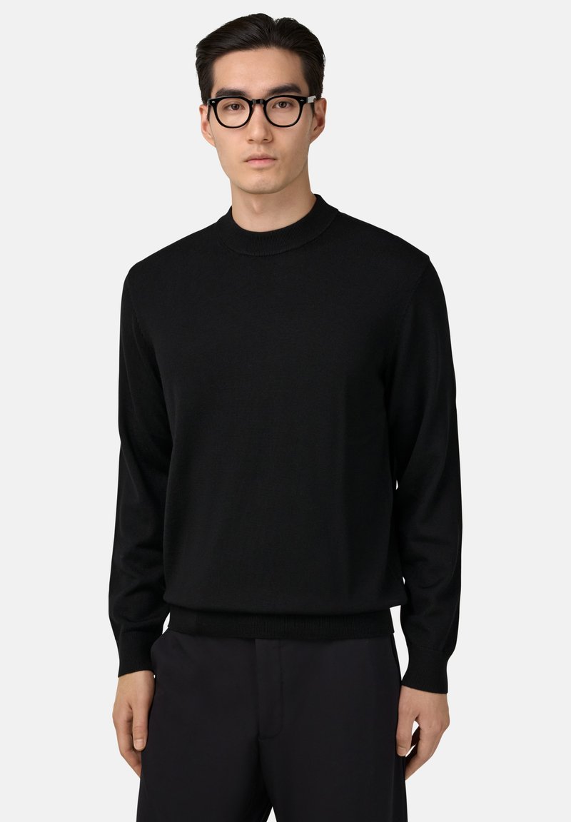 Pull en maille noir avec un col haut, des manches longues et des poignets et un ourlet côtelés. Texture lisse, design ajusté. Porté avec un pantalon sombre.