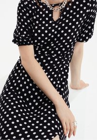 Robe noire à pois blancs, manches courtes bouffantes, décolleté en forme de clé, et taille cintrée. Tissu lisse avec un léger éclat.