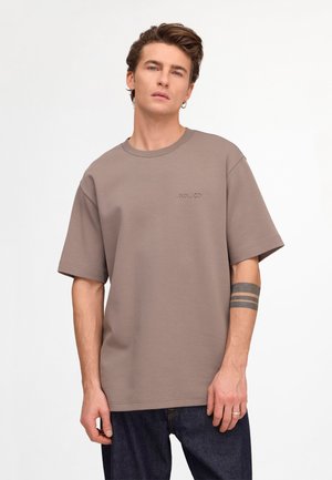 Ung mand med kort brunt hår bærer en taupe oversized t-shirt med "NN.07" broderi, mørke jeans og en sort stribet armtatovering, står stille.