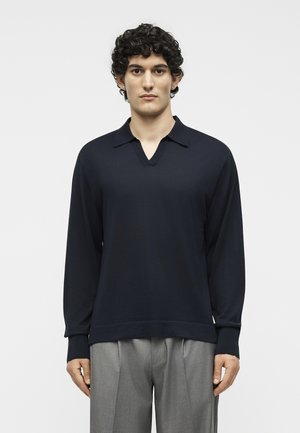 Νέος άνδρας με σγουρά μαλλιά φοράει μαύρο πουκάμισο με μακριά μανίκια και γιακά και γκρι πλισέ παντελόνια, στέκεται σε απλό ανοιχτό φόντο.