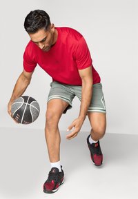 Κόκκινο αθλητικό T-shirt με μαύρο λογότυπο, γκρι σορτσάκι με πλάγιες ρίγες, μαύρα και κόκκινα παπούτσια. Παίκτης ντριμπλάρει μια μπάλα καλαθοσφαίρισης.