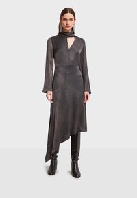 Čierne šaty s dlhým rukávom, vysokým golierom a vykrojením v tvare kľúča. Hladká, textúrovaná látka, asymetrický lem, vrstvený dizajn. Doplnkové čierne čižmy s hladkým vzhľadom.