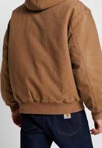 Brun huppetröja med ribbad nederkant och mudd, med en strukturerad yta. Bakifrån visas en logotypetikett på mörka jeans i denim.