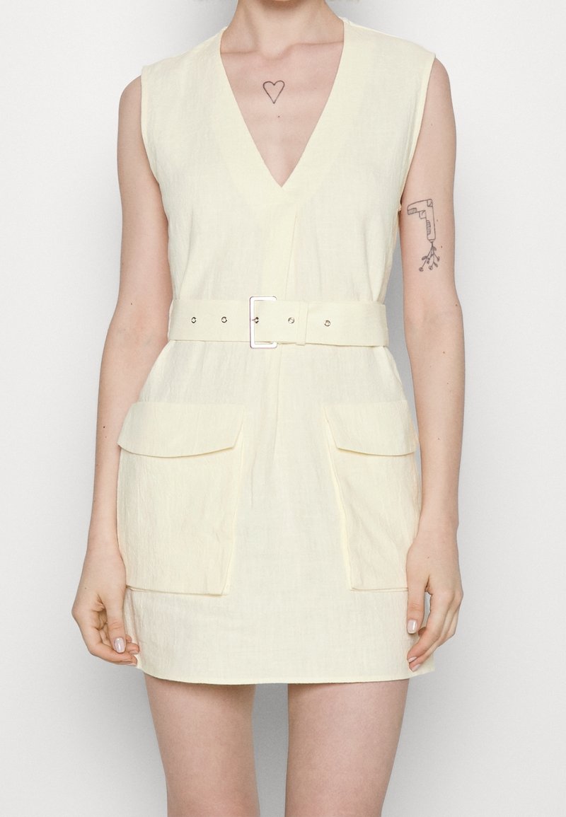 Robe sans manches jaune pâle avec un décolleté en V profond, taille ceinturée et deux poches avant. Tissu texturé avec un aspect légèrement froissé.