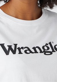 Bílý bavlněný tričko s kulatým výstřihem, na kterém je výrazné černé logo "Wrangle" potisknuté přes hruď.