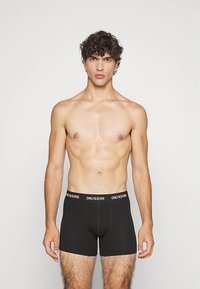 Moški brez majice s kodrastimi lasmi, oblečen v črne bokserice ONLY&SONS, stoji obrnjeno naprej proti enobarvni svetli podlagi.