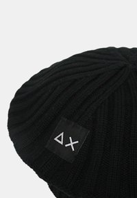 Berretto lavorato a maglia nero con trama a coste, con una toppa quadrata con il logo con le lettere "ΔX" in argento. Profilo superiore arrotondato.